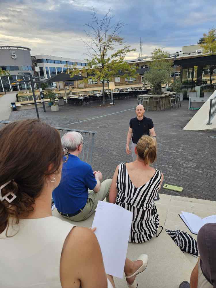 Een spreker oefent buiten op de Corda Campus terwijl een kleine groep leden op de trappen toekijkt.