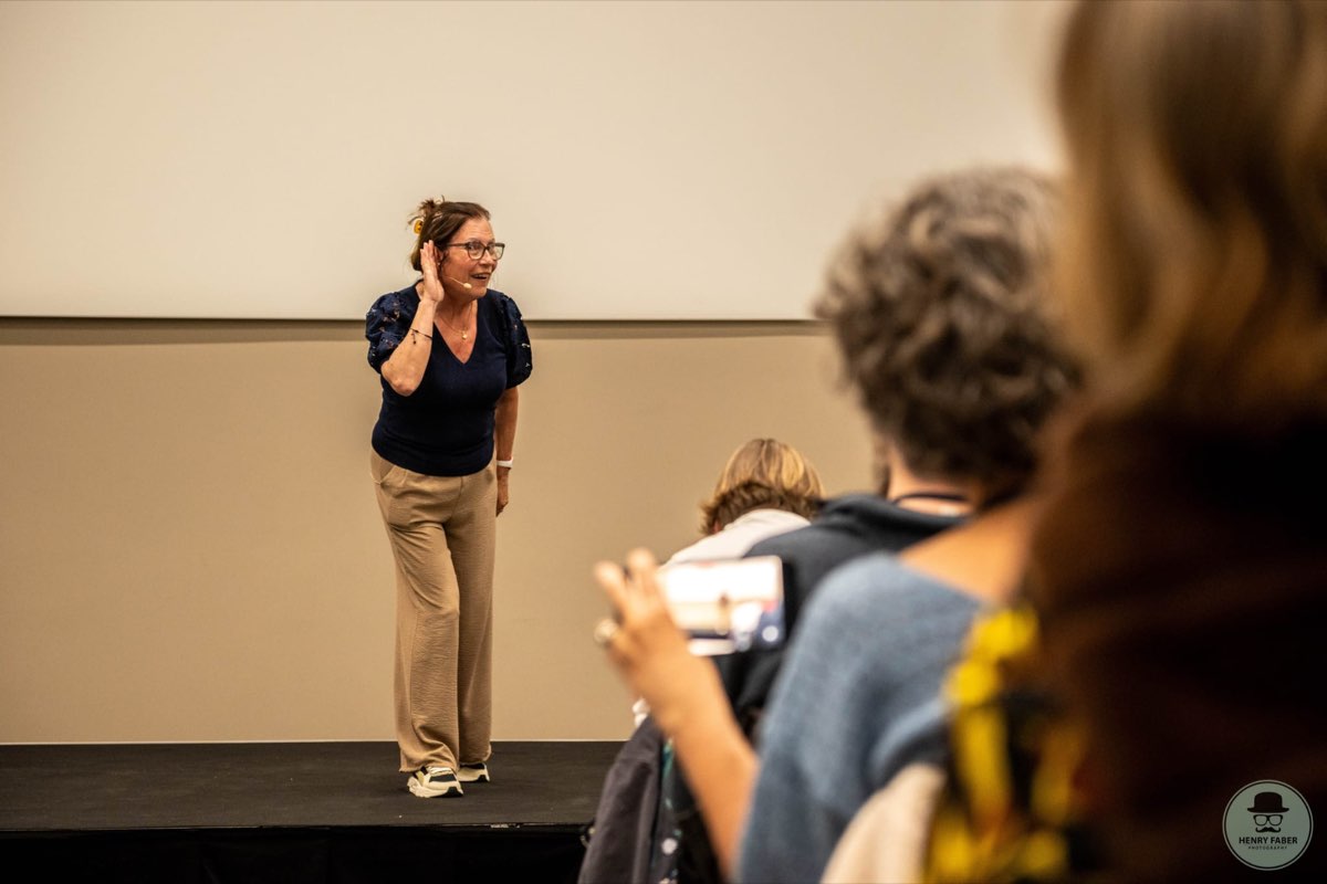 Een spreker op het podium bij Toastmasters Hasselt houdt een hand achter haar oor terwijl het publiek toekijkt.